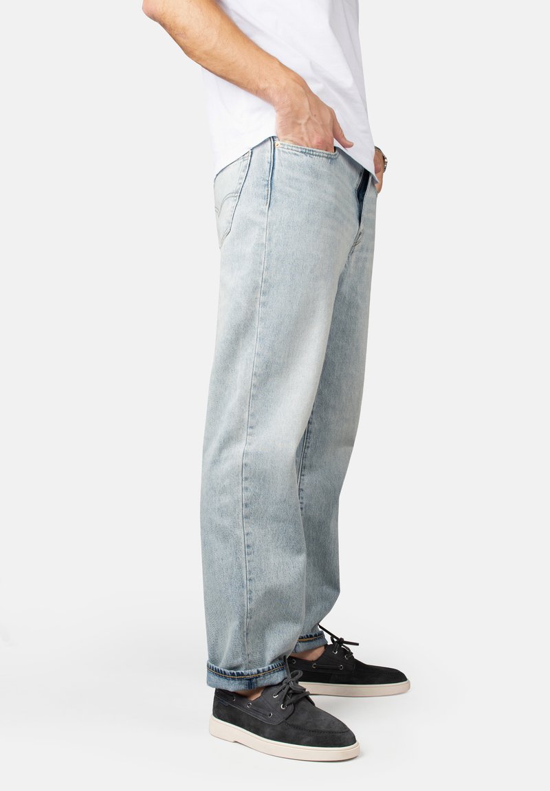 Lichtblauwe denim jeans met een ontspannen pasvorm, voorzien van riemlussen en zijzakken. Gecombineerd met donkere suède schoenen en een wit shirt.