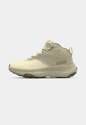 Hoka Wanderschuh in hellbeige mit strukturiertem Obermaterial, Schnürverschluss und Vibram-Sohle. Auffälliges Seitenlogo und unterstützendes Design.