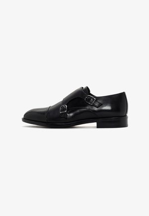 Scarpa formale in pelle nera con doppia fibbia monk strap, tacco basso e punta arrotondata, esposta su uno sfondo bianco.