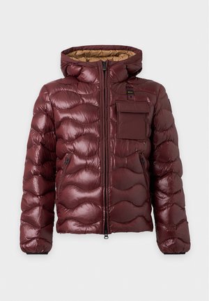 Jeune homme portant une veste matelassée bordeaux avec capuche, chemise noire, jean clair à jambes larges et baskets blanches sur fond blanc uni.