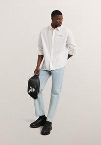 Camisa blanca de manga larga con cuello abotonado, jeans azul claro, zapatillas negras y una bolsa negra con diseño de personaje de dibujos animados.