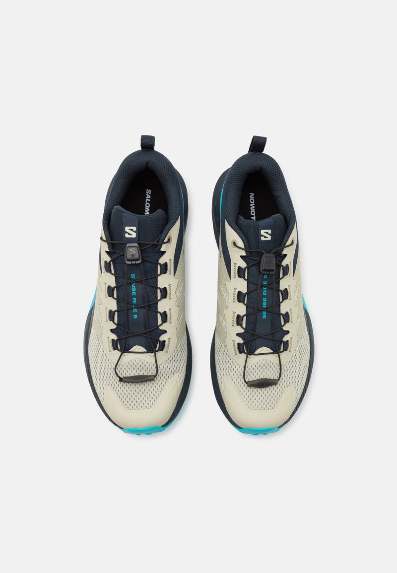 Zapatillas deportivas de color crema claro y navy con parte superior de malla, acentos turquesa y una suela robusta; cuenta con un sistema de cordones rápidos y un collar acolchado.