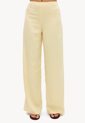 Pantalon jaune pâle taille haute à jambes larges porté avec des sandales ouvertes foncées, présenté de la taille aux pieds sur un fond uni.