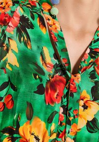 Florale Bluse mit lebhaft grünem Hintergrund und kräftigen roten, orangefarbenen und gelben Blumen sowie einem V-förmigen Ausschnitt. Weiche, glatte Textur.