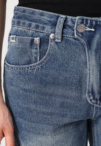Svijetloplave traperice imaju visok struk, kopčanje na dugme i patentni zatvarač, te dva prednja džepa sa šavom. Imaju sužene nogavice.