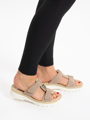 Piedi con sandali beige in camoscio con fibbie decorative e suole bianche, abbinati a leggings neri su sfondo bianco.