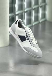 Weißer und grauer Sneaker mit einer Mischung aus glattem Synthetik- und Wildledermaterial, mit einem dunkelblauen Akzent und weißen Schnürsenkeln, in der Luft schwebend dargestellt.