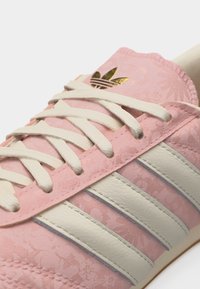 Ροζ αθλητικό παπούτσι με λευκές δερμάτινες ρίγες, λευκά κορδόνια, διακριτική λουλουδένια υφή και χρυσό λογότυπο Adidas trefoil στη γλώσσα.