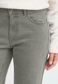 Mujer vestida con jeans de tiro alto gris y un suéter de punto rosa claro, con la mano relajada a su lado.