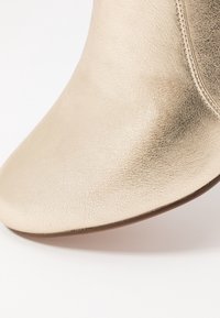 Metalická zlatá kotníková bota s hladkým, členitým koženým povrchem, špičatou špičkou a jemným stehováním podél švu.