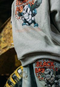 Grijze trui met kleurrijke gorillagrafiek en tekst op de voorkant; blauwe spijkerbroek met ingewikkeld oranje borduursel op het achterzakje.