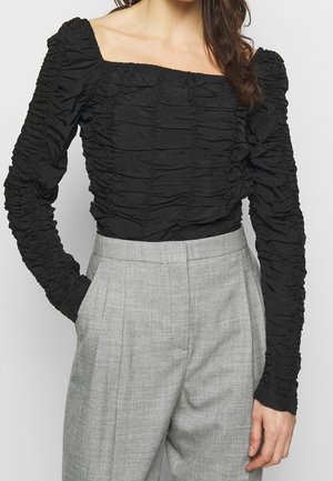 Vrouw draagt een zwart gerimpeld shirt met lange mouwen en een vierkante halslijn, gecombineerd met hoge taille grijze plissébroek, handen in de zakken.