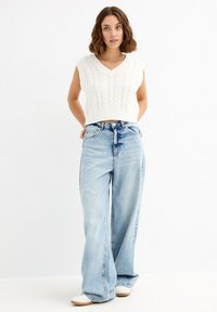 Joven mujer con cabello corto y ondulado, vistiendo un suéter sin mangas de punto blanco, jeans de pierna ancha de color azul claro y zapatillas blancas.