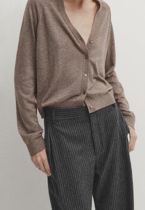 Persoon die een bruine gebreide cardigan met knopen draagt en een donkergrijze broek met fijne krijtstrepen en hoge taille, met handen bij de zakken, tegen een effen achtergrond.