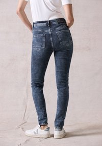 Vaqueros de mezclilla oscuros y ajustados, con cinco bolsillos y un ligero desvanecimiento. Combinados con zapatillas blancas con acentos grises, vista desde atrás.