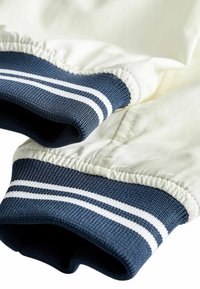 Poignets élastiques avec tricot côtelé rayé bleu et blanc sur les manches d'une veste de couleur claire, montrant la texture du tissu et les détails des coutures.