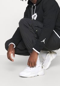 Sneakers atletiche bianche con lati texturizzati, lacci neri e branding "ZOOM". Indossati con joggers neri e una felpa nera con dettagli del logo.