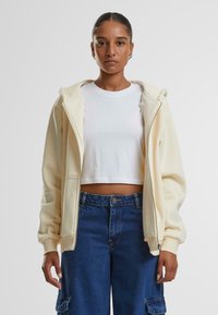 Lichtromige zip-up hoodie met een zachte textuur, met een kangoeroezak, gedragen over een witte crop top en gecombineerd met blauwe denim cargo pants.