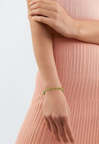 Goldkette-Armband an einem Handgelenk, bestehend aus verbundenen Metallsegmenten mit glänzender Oberfläche. Die Hand ruht auf einem strukturierten rosafarbenen Kleidungsstück.