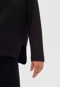 Felpa nera con maniche lunghe, vestibilità rilassata, fessure laterali e texture morbida. Mano appoggiata di lato, che mette in evidenza il design semplice.