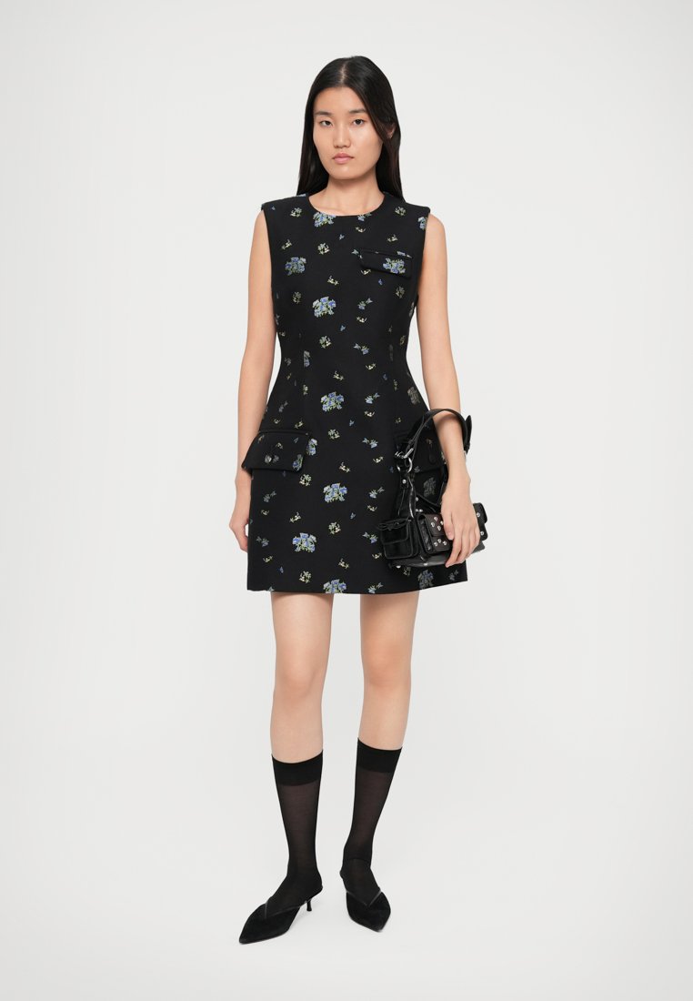 Robe noire sans manches avec imprimé floral, deux poches et un ourlet évasé. Associée à des chaussettes noires montantes jusqu'aux genoux et des ballerines pointues.