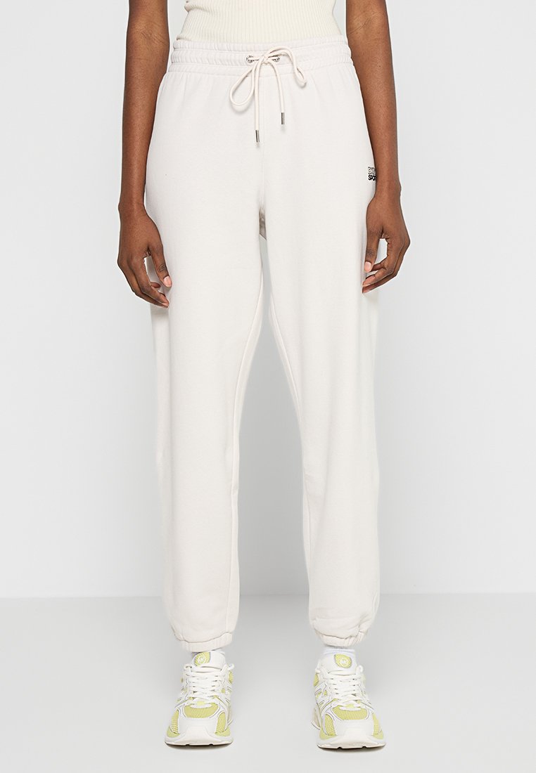 Jasno beżowe dresy ze sznurkiem w talii, elastycznymi mankietami i subtelnym detalem logo. Miękka tkanina o luźnym kroju dla komfortu.