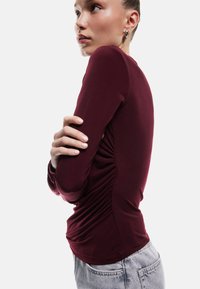 Bordeaux langemouwen top met verzamelde zijdetails, een gladde textuur en een aansluitende silhouet, gecombineerd met lichtblauwe jeans.