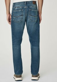 Blauwe denim jeans met een recht gesneden pijp, voorzien van twee achterzakken en lichte wassing, tegen een neutrale achtergrond.
