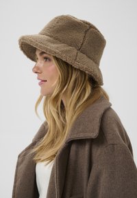 Rjav klobuk s širokim obodom, izdelan iz puhaste tkanine, z urejenim oblikovanjem. Nosijo ga z usklajenim rjavim plaščem in belim zgornjim delom.