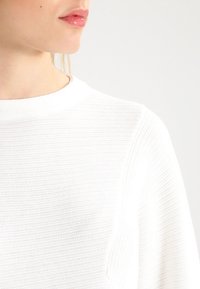 Vit stickad tröja med rund halsringning, med texturerade ränder, långa ärmar och en figursydd silhuett.