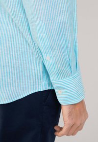 Camisa de rayas azul claro y blanca, hecha de tejido ligero. Presenta un puño abotonado y una textura suave con un diseño ligeramente ajustado.
