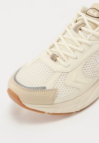 Zapatilla deportiva de color crema con parte superior de malla transpirable, acentos de cuero suave, suela de goma y sutiles detalles en gris y beige.