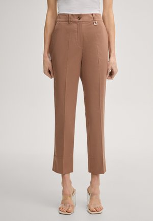 Kobieta w brązowych spodniach o podwyższonym stanie, skróconych, z zakładkami z przodu, brązowym guzikiem oraz sandałach na słupku z przezroczystym paskiem.