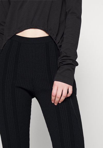 Blusa preta de mangas compridas com comprimento encurtado e detalhes franzidos, combinada com calças pretas caneladas de cintura alta com riscas verticais.