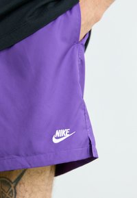 Fialové šortky Nike, ľahká látka, bočné vrecko, biele logo na leme, bočné rázpory pre flexibilitu, hladká textúra.