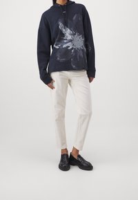 Marinblå hoodie med ett stort blommönster i grått, kombinerat med vita byxor och svarta chunky loafers. Tyget verkar mjukt och avslappnat.