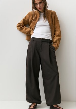 Frau trägt eine hellbraune Wildlederjacke über einem weißen Oberteil, dunkle, weit geschnittene Nadelstreifenhose und schwarze Schuhe, steht vor einer schlichten weißen Wand.