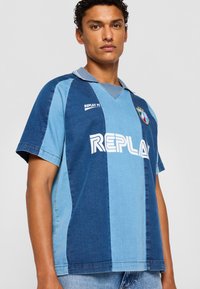 Džinsinis futbolo marškinėlis įvairių atspalvių mėlyna spalva, su trumpomis rankovėmis, apykakle ir ryškiu baltu "REPLAY" logotipu priekyje.
