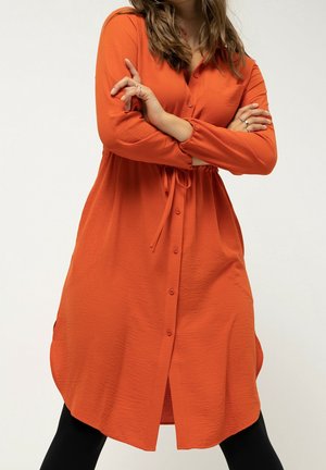Femme portant une robe orange à manches longues boutonnée avec une ceinture à la taille et des leggings noirs, les bras croisés sur la poitrine.