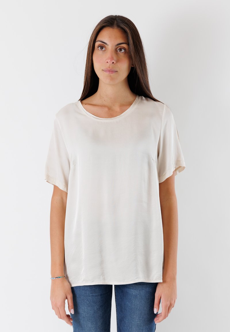 Top corto color beige chiaro realizzato in tessuto liscio con scollatura rotonda. Presenta un fit rilassato e una caduta delicata. Indossato con jeans blu.