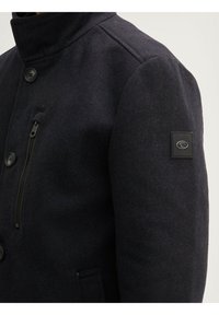 Chaqueta de mezcla de lana en azul marino con cuello alto, con grandes botones, un bolsillo con cremallera y un parche de marca en la manga izquierda.