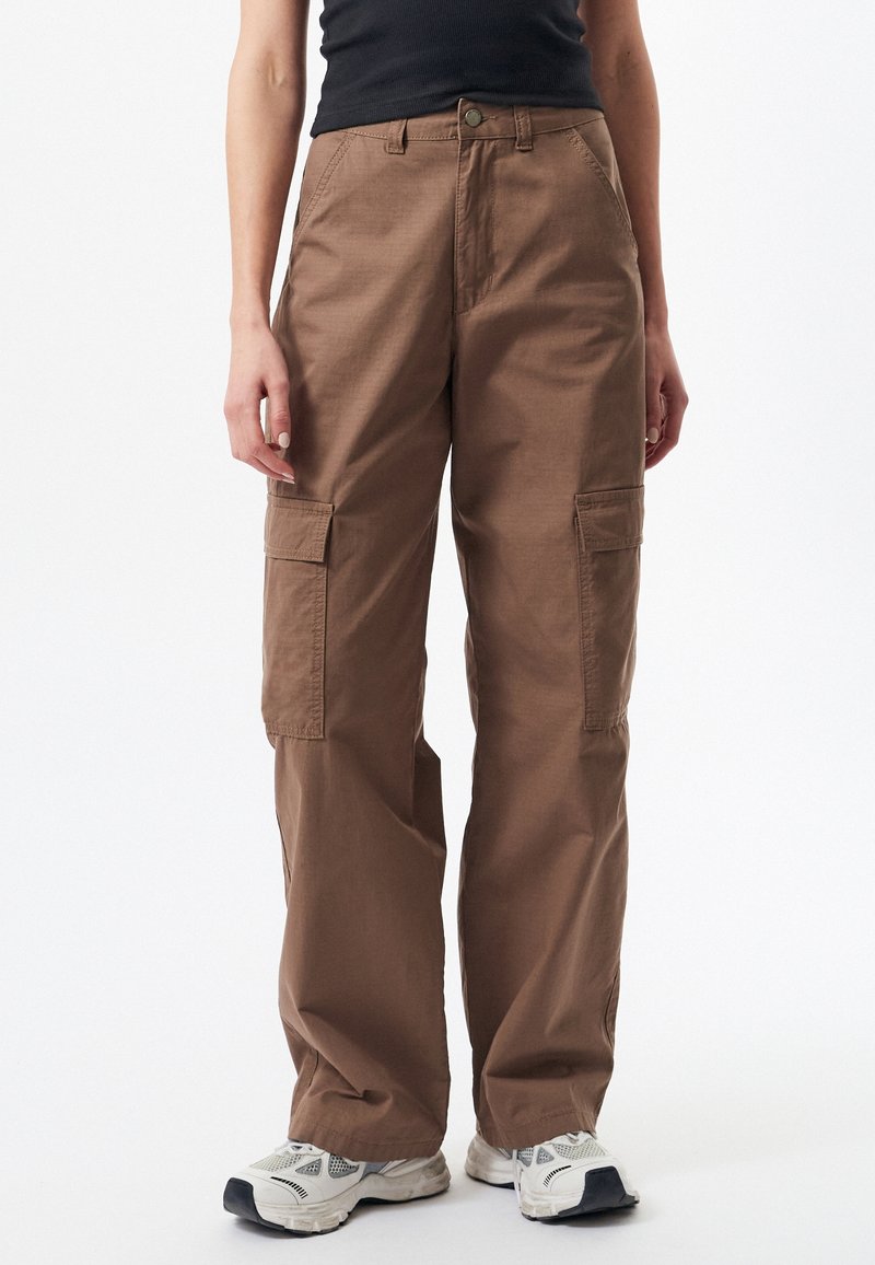 Pantalon cargo marron à jambes larges avec poches à rabat à l'avant, porté avec des baskets blanches et un haut sans manches noir.