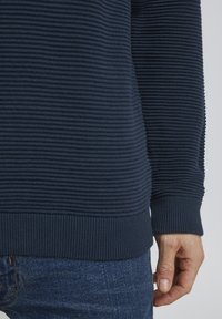 Marineblau gestrickter Pullover mit horizontaler Rippstruktur, ausgestattet mit einem engen Bündchen und Saum, getragen über blauen Denim-Jeans. Detaillierte Nahaufnahme des Ärmels und der Hand.