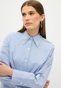 Camicia a righe blu chiaro con colletto a punta, bottoni bianchi e maniche lunghe. Il tessuto appare liscio con una leggera lucentezza.