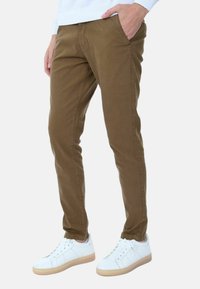Pantaloni di cotone marroni con vestibilità slim, tasche anteriori e una texture liscia, abbinati a sneaker bianche con suola di gomma chiara.