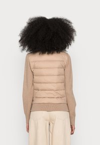 Beige puffer vest met een gequilte structuur, gecombineerd met een tan breisel trui. Zachte textuur, ronde hals en geribde tailleband. Casual pasvorm.