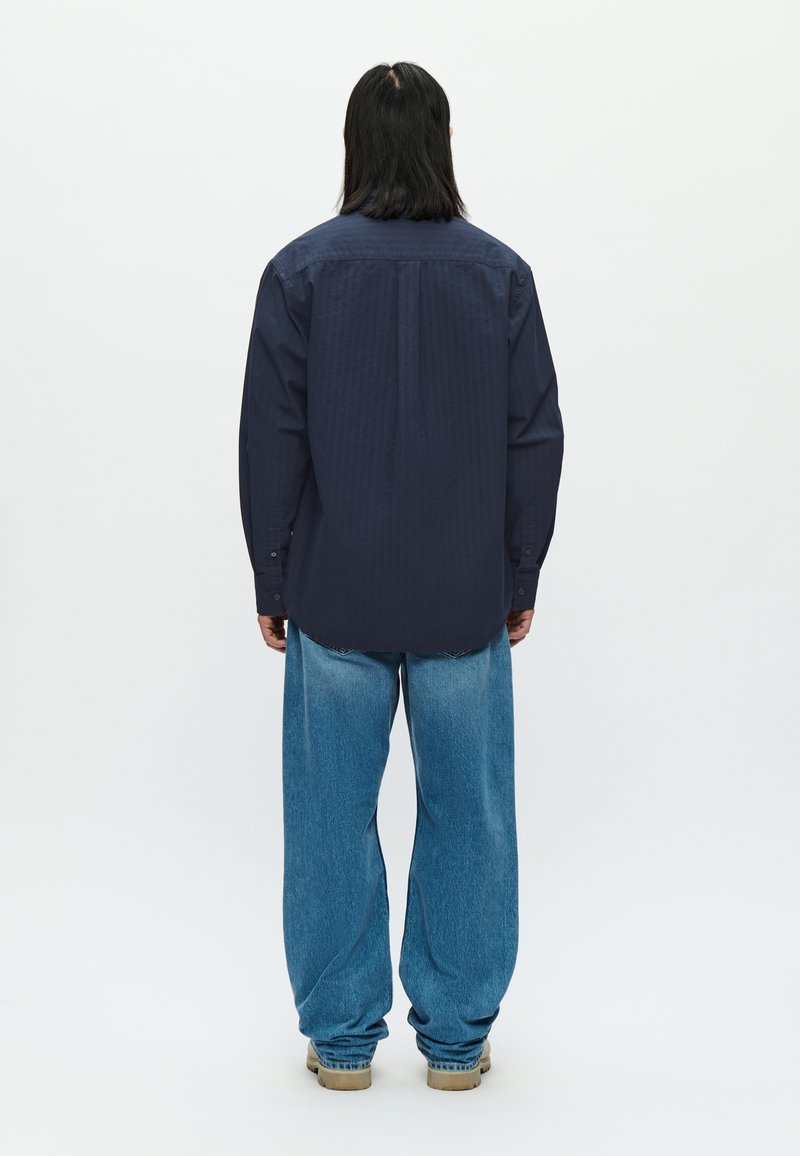 Marineblaues Hemd mit Knöpfen, vertikaler Rippstruktur, lockerer Passform und abgerundetem Saum, kombiniert mit weit geschnittenen, hellblauen Jeans und beigem Schuhwerk.