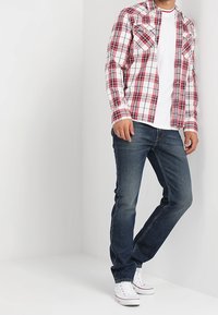 Camicia a maniche lunghe a quadri rossi e bianchi sopra una maglietta bianca, abbinata a jeans blu scuro e sneakers bianche.