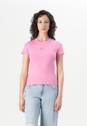 Camiseta rosa de manga corta hecha de algodón suave. Presenta un detalle de corte en forma de corazón en el escote. Combinada con jeans de mezclilla rasgados de color azul claro.