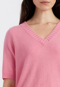 Maglione corto a maniche corte di colore rosa chiaro con design a V con righe. Tessuto morbido, vestibilità leggermente ampia, dettagli lungo il collo e le maniche.